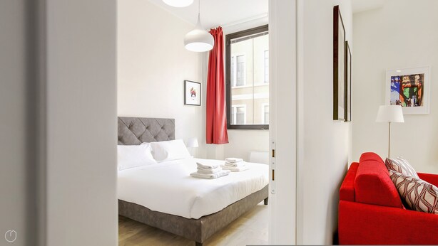 Una habitación de hotel con una cama, un sofá rojo, una ventana con cortinas, un cuadro enmarcado y una lámpara.