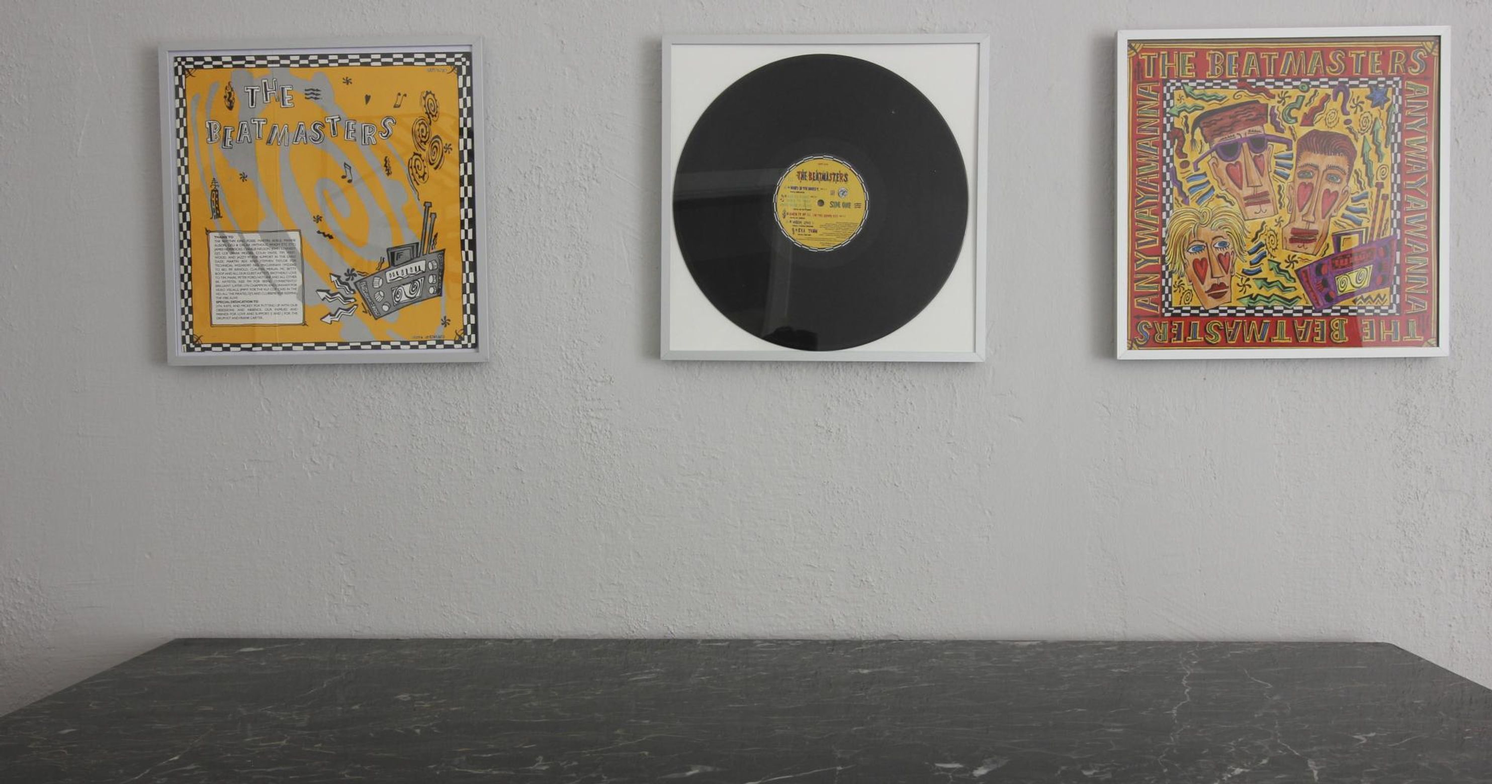 Three framed items on a wall: a yellow poster with text and graphics, a black vinyl record, and a colorful poster with text and graphics.
