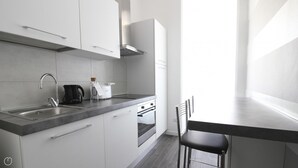 Une cuisine moderne avec des meubles blancs, un évier en acier inoxydable, une bouilloire, un grille-pain et une cuisinière. Une fenêtre laisse entrer la lumière naturelle et une chaise est placée à la table.