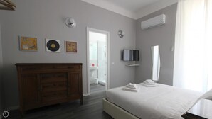 Une chambre à coucher moderne, avec une commode en bois, un lit recouvert de linge de maison blanc, un téléviseur à écran plat et des œuvres d’art accrochées aux murs.