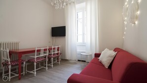 A room with a red sofa, a wooden dining table with chairs, a TV mounted on the wall, and a radiator. - Italianway Easy - Melzo 12 - Topolino (Milan)