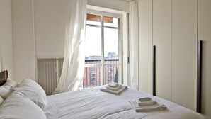 A hotel room with a large bed, white linens, a window with sheer curtains, and a view of buildings. - Mi-navp112a6 - Naviglio Pavese 112 (Milan)