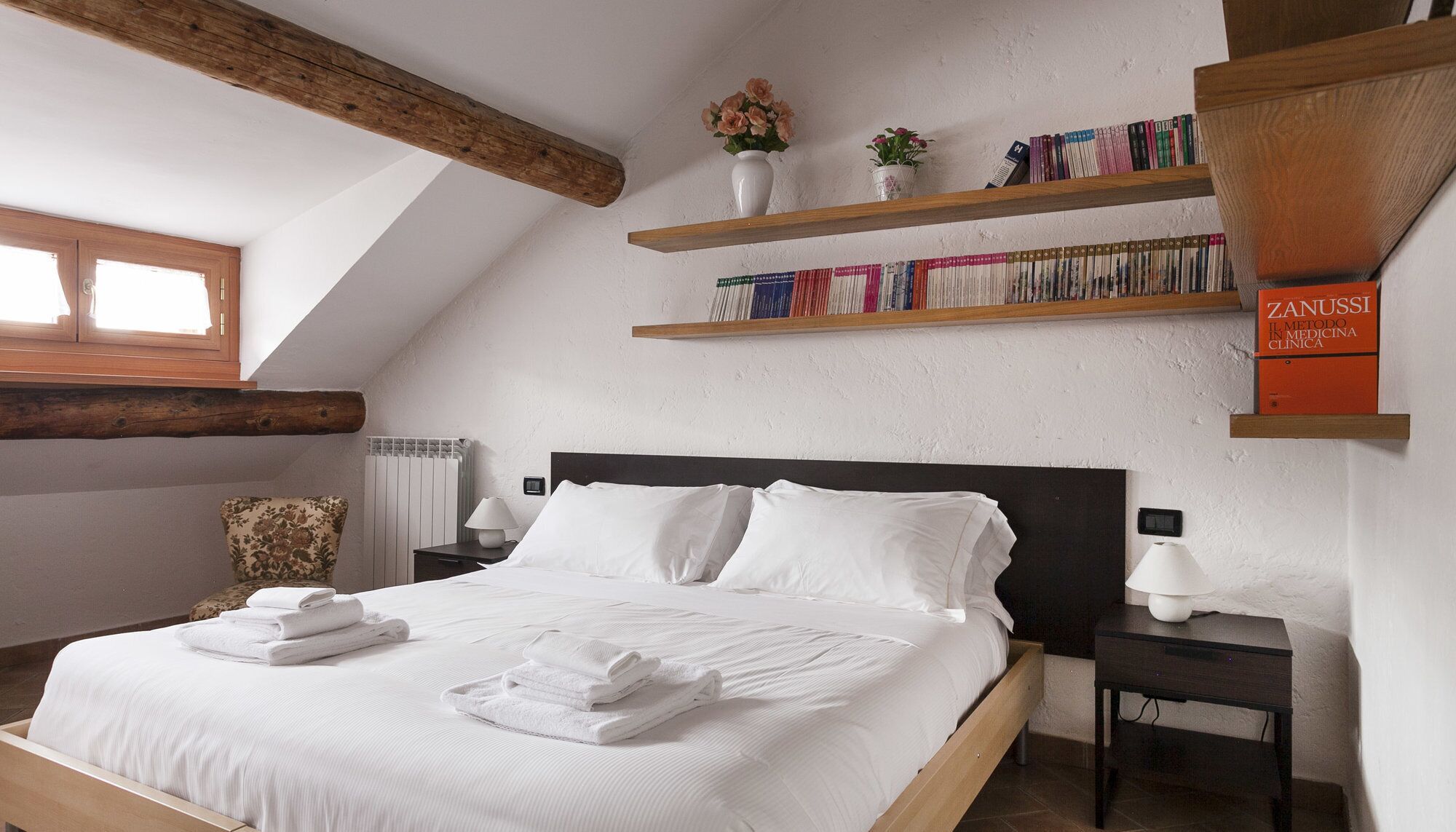 A bedroom with a wooden ceiling, a bed with white linens, a nightstand with a lamp, a bookshelf with books, and a window with curtains.