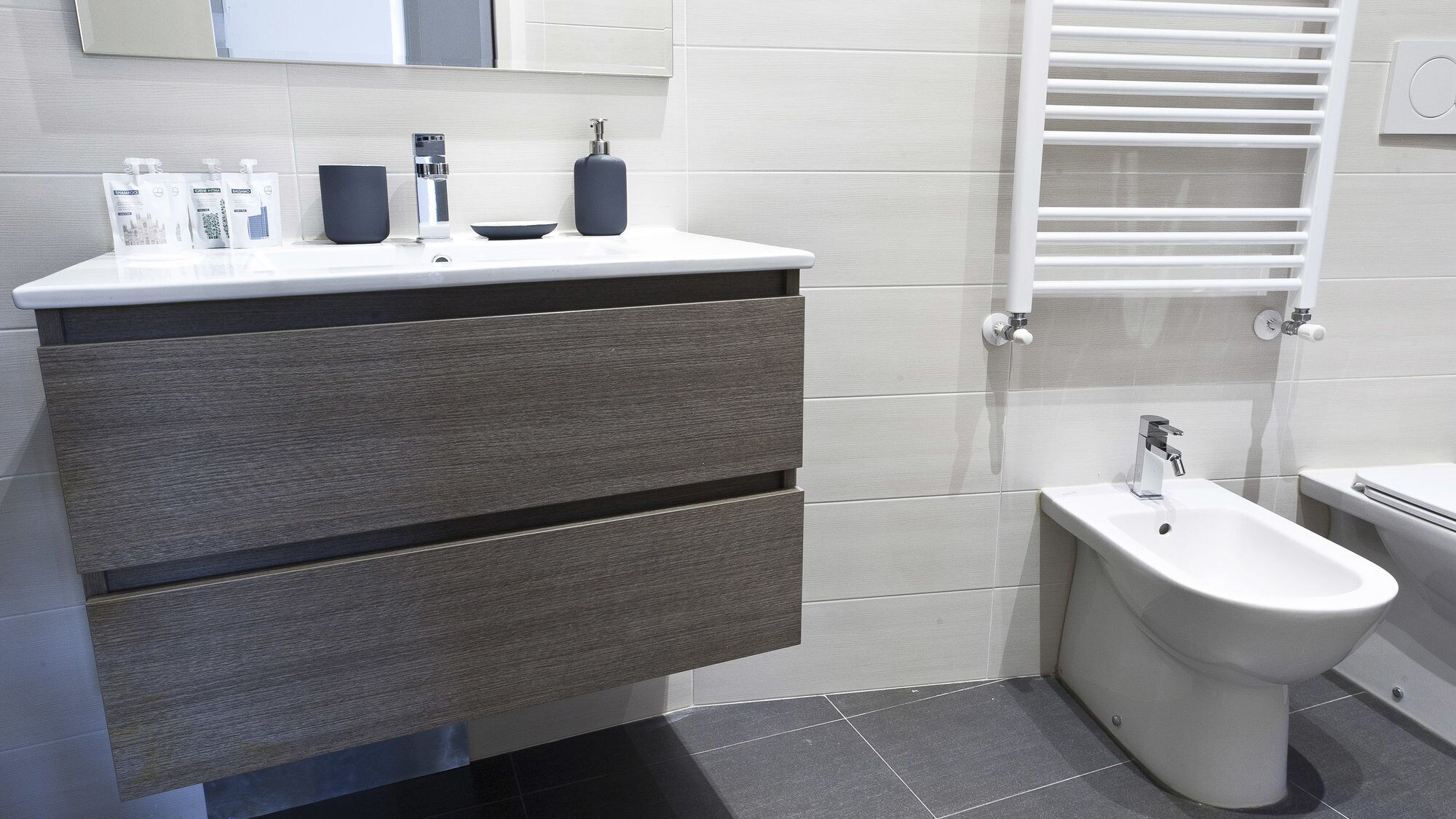 Un baño moderno con un mueble de tocador gris, un bidé y un perchero blanco.