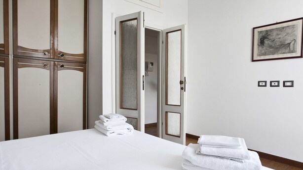 A hotel room with a bed, a stack of towels, a wardrobe, and a framed artwork on the wall. - Italianway Easy - Piazza Firenze 4 (Milan)