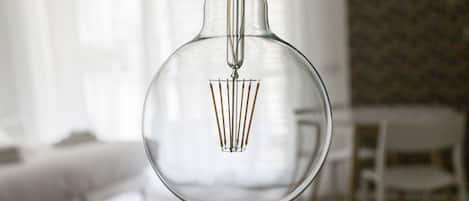 A clear glass light bulb with a visible filament and a metal base, hanging in a room with a blurred background.