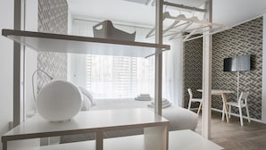 A modern hotel room with a minimalist design, featuring a bed, a white table with a spherical object, a hanging clothes rack, and a TV mounted on a geometric-patterned wall.
