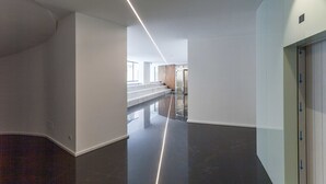 A modern, spacious hallway with a glossy black floor, white walls, and a glass elevator.