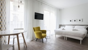 Quarto de hotel com uma cama, uma poltrona amarela, uma mesa pequena e uma televisão fixada na parede.