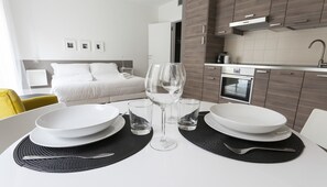 Una moderna camera d'hotel con angolo ristoro, che include un tavolo bianco apparecchiato con piatti, bicchieri e un calice da vino. La stanza dispone anche di un letto, un piccolo angolo cottura con microonde e forno, e un bagno visibile sullo sfondo.