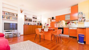 Uma cozinha moderna com armários laranjas, uma área de refeições com duas cadeiras e uma sala de estar com televisão e estantes.