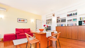 Uma sala de estar moderna com um sofá vermelho, uma mesa de jantar de madeira com duas cadeiras, uma estante repleta de livros e uma televisão.