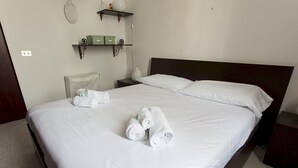 Une chambre d’hôtel avec un grand lit, du linge de lit blanc, deux serviettes enroulées, une table de chevet, une petite table et une étagère avec des boîtes vertes et un vase.