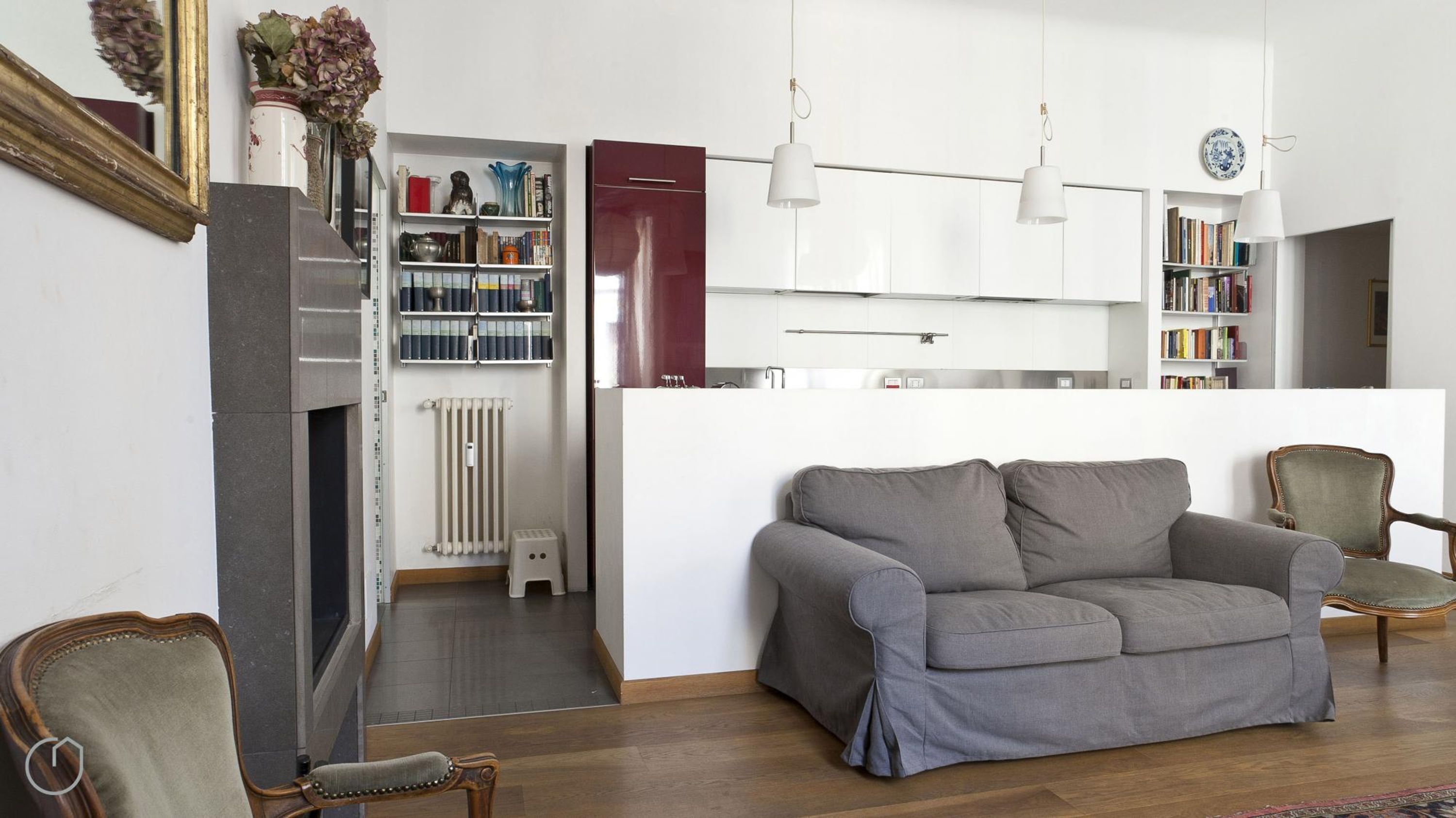Ein modernes Wohnzimmer mit grauem Sofa, Holzstühlen, einem Bücherregal und einer Küchenzeile mit weißen Schränken sowie einem Heizkörper.
