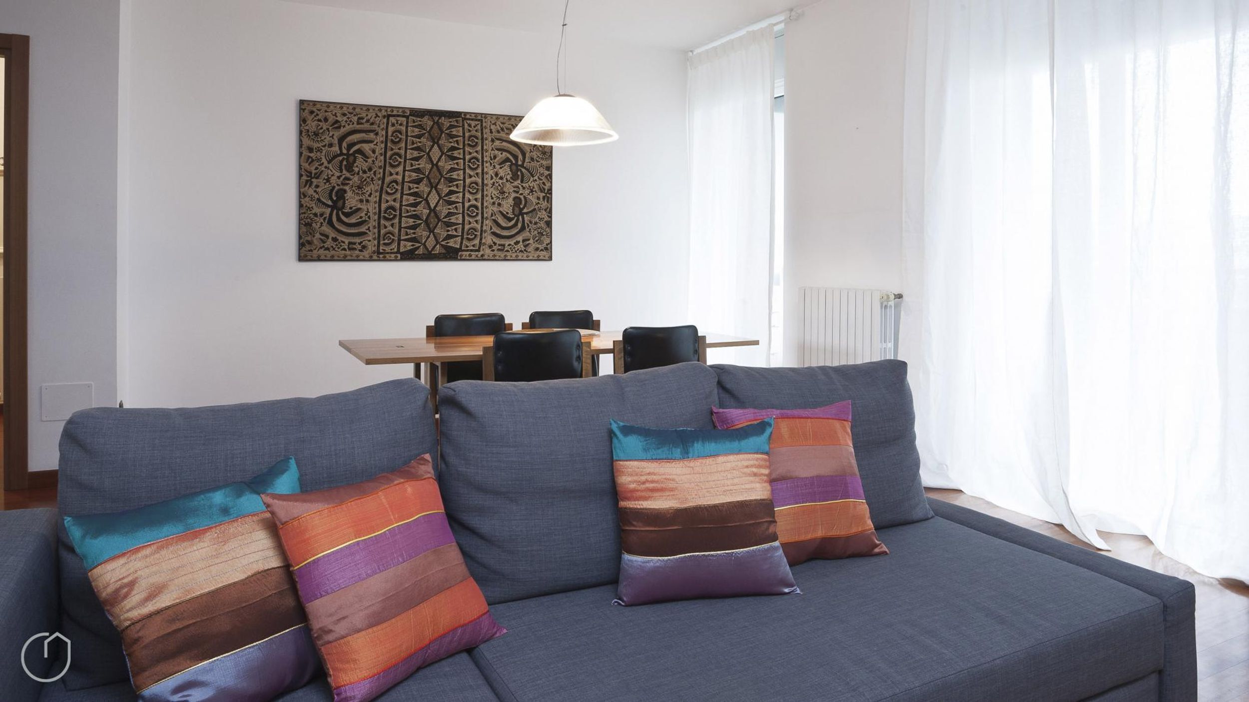 A modern living room with a grey sofa, colorful striped cushions, a dining table with chairs, and a pendant light.