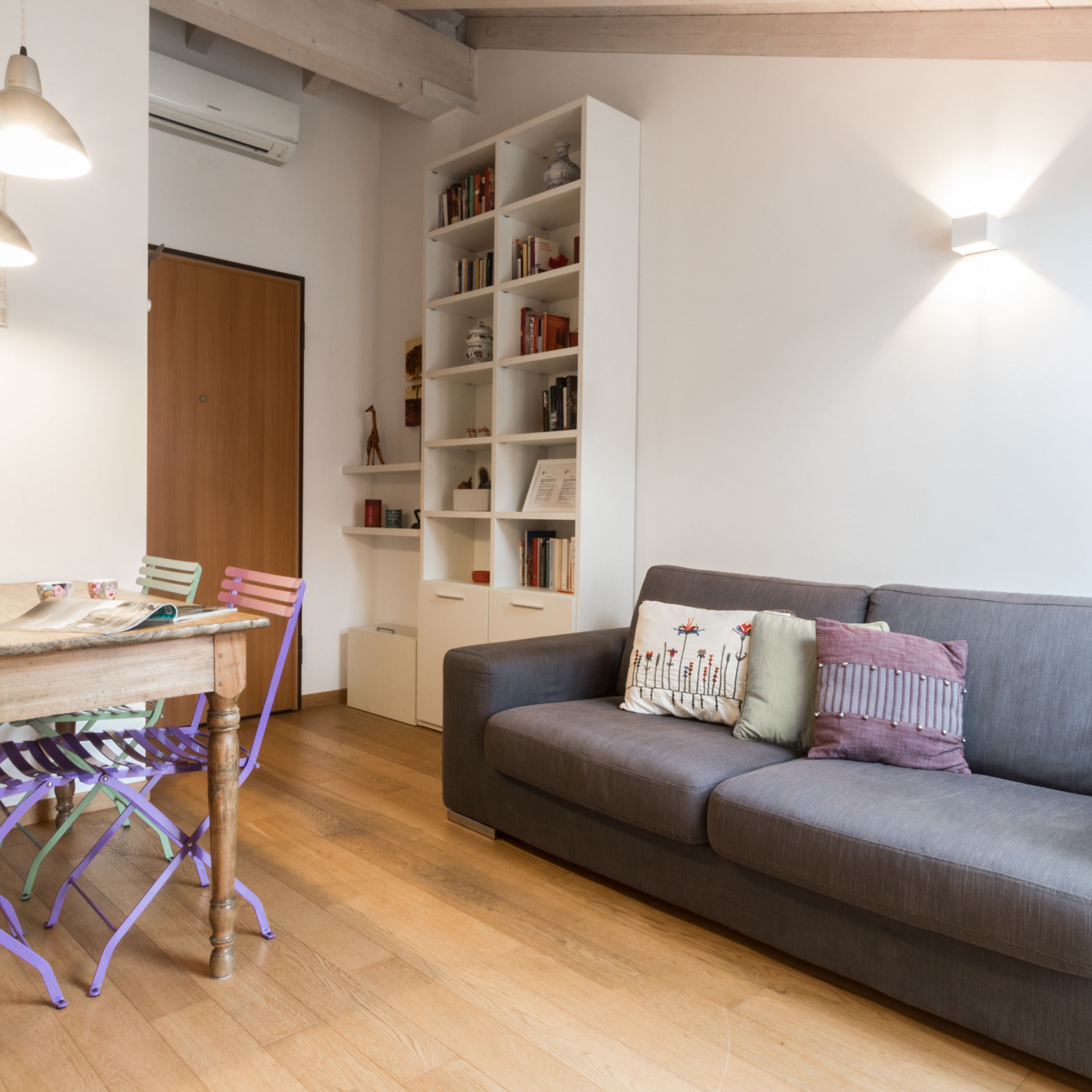 A living room with a grey sofa, a wooden table with chairs, a bookshelf, and a door.