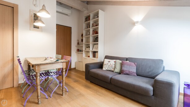 A cozy living room with a grey sofa, a wooden dining table with colorful chairs, and a white bookshelf filled with books and decorative items. - Italianway Easy - Savona 108 (Milan)
