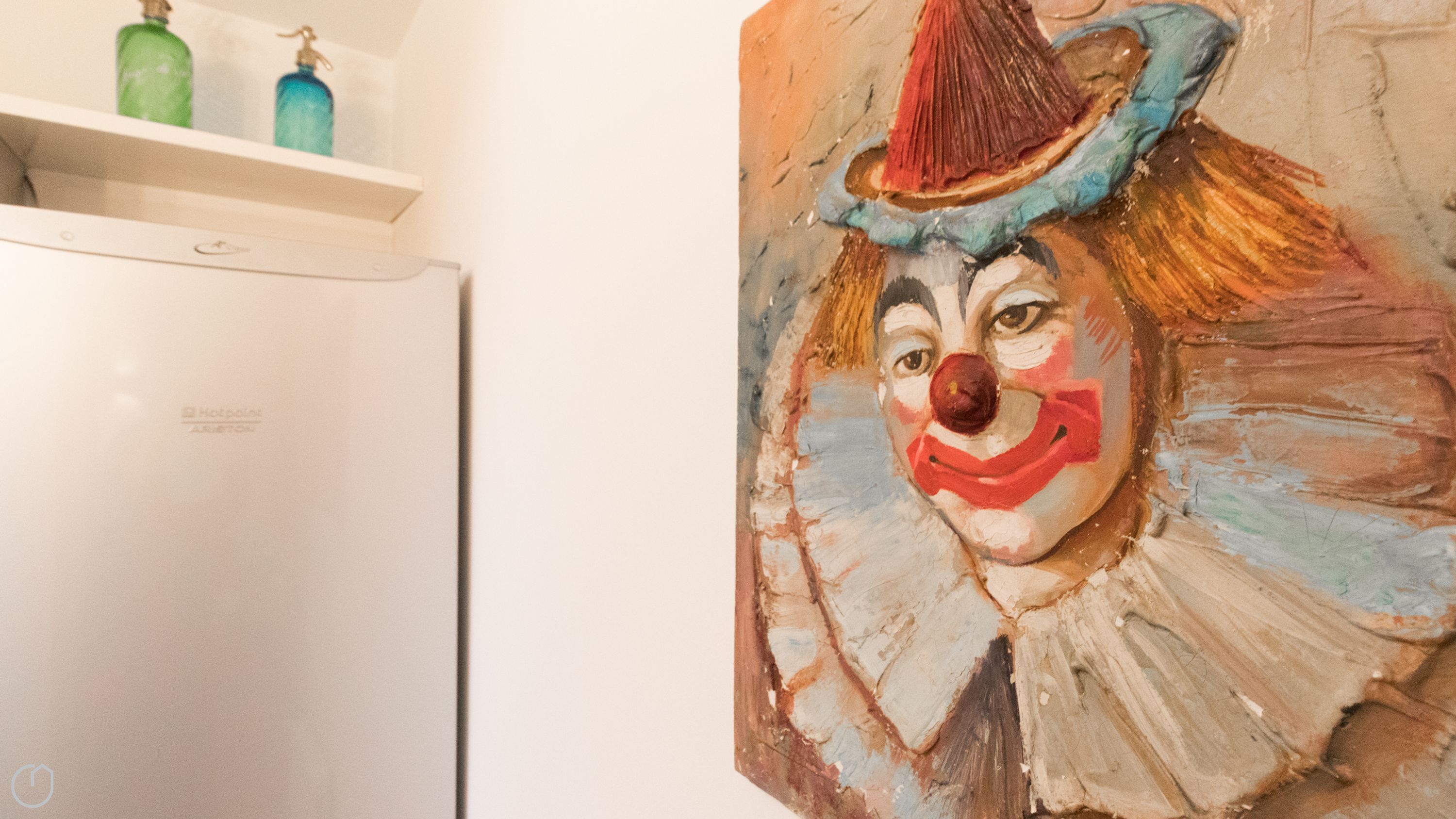 A white refrigerator with a Hotpoint logo, a wall with a colorful clown painting, and a shelf with two green bottles.