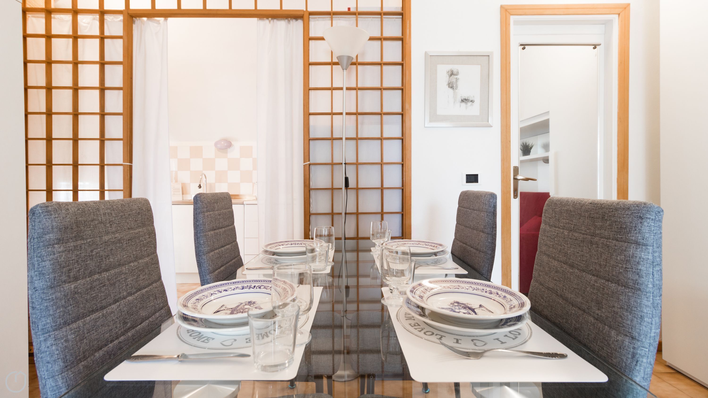 A dining area with a glass table set for four, grey upholstered chairs, and a view of a kitchen and a door in the background.