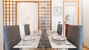 Un comedor con una mesa de vidrio dispuesta para cuatro, sillas grises tapizadas y vistas a una cocina y una puerta al fondo.