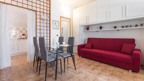 A modern living room with a red sofa, a glass coffee table, and grey chairs. There is a kitchen area with white cabinets and a white countertop. - Washington 91 (Milan)