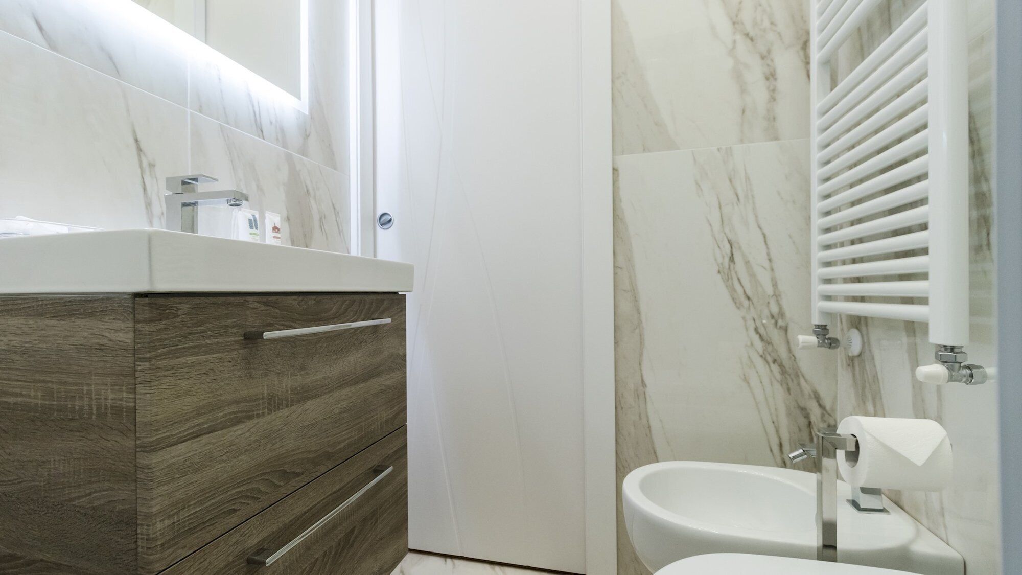 A modern bathroom with a marble wall, a wooden vanity, a toilet, and a bidet.