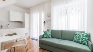 A modern living room with a green sofa, white curtains, and a kitchen area with white cabinets. - Italianway Easy - Ciro Menotti 18 (Milan)