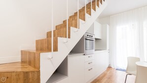 A modern kitchen with white cabinets and a built-in oven, a wooden staircase, and a dining area with a white chair. - Italianway Easy - Ciro Menotti 18 (Milan)