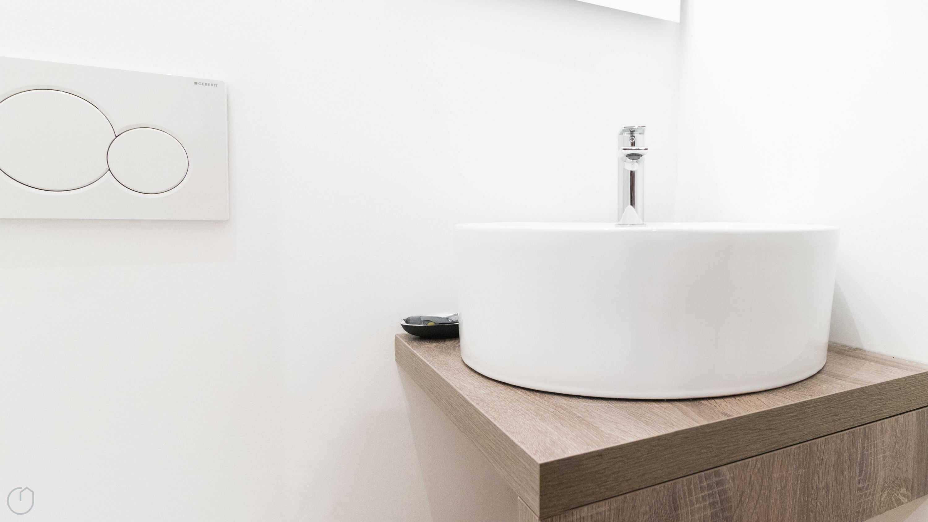 A modern bathroom with a round white sink on a wooden countertop, a wall-mounted light switch, and a small black object on the counter.