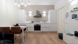 A modern kitchen with white cabinetry, a built-in oven, and a stainless steel range hood. There is a dining area with a white table and brown chairs. - Mi-vige13a3 - Vigevano 13 (Milan)