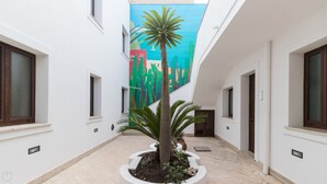 A narrow corridor with white walls, a central palm tree, and a colorful mural on the back wall. - Ag-e431-bonf8f1 Ottoventi Apartments Tramontana (Lampedusa)