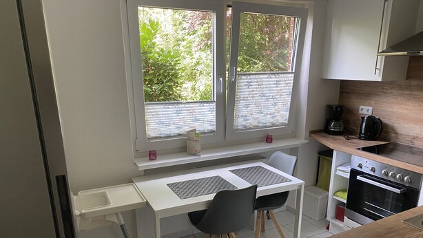 Een compacte keuken met een wit aanrecht, een inbouwoven en een raam met zonwering.