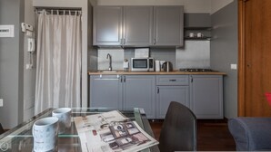 A compact kitchen with grey cabinets, a microwave, a kettle, and a stove. There's a glass table with a newspaper and a chair. - Italianway Easy - Paisiello 4 A (Milan)