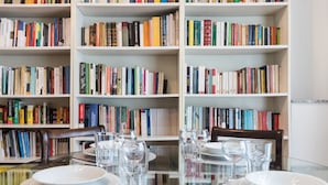 Un comedor con una mesa de vidrio rodeada de sillas, una estantería al fondo y algunos vasos de vino vacíos sobre la mesa.