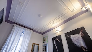 A room with a high ceiling, decorative moldings, a large window with white curtains, and two framed photographs of dancers.