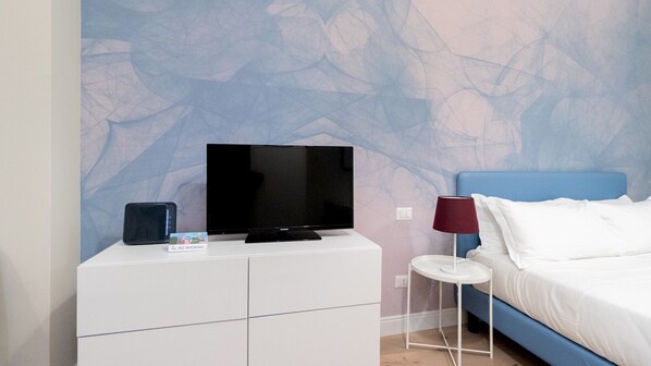 A hotel room with a bed, a TV on a white dresser, a small table with a lamp, and a wall with a leaf pattern.