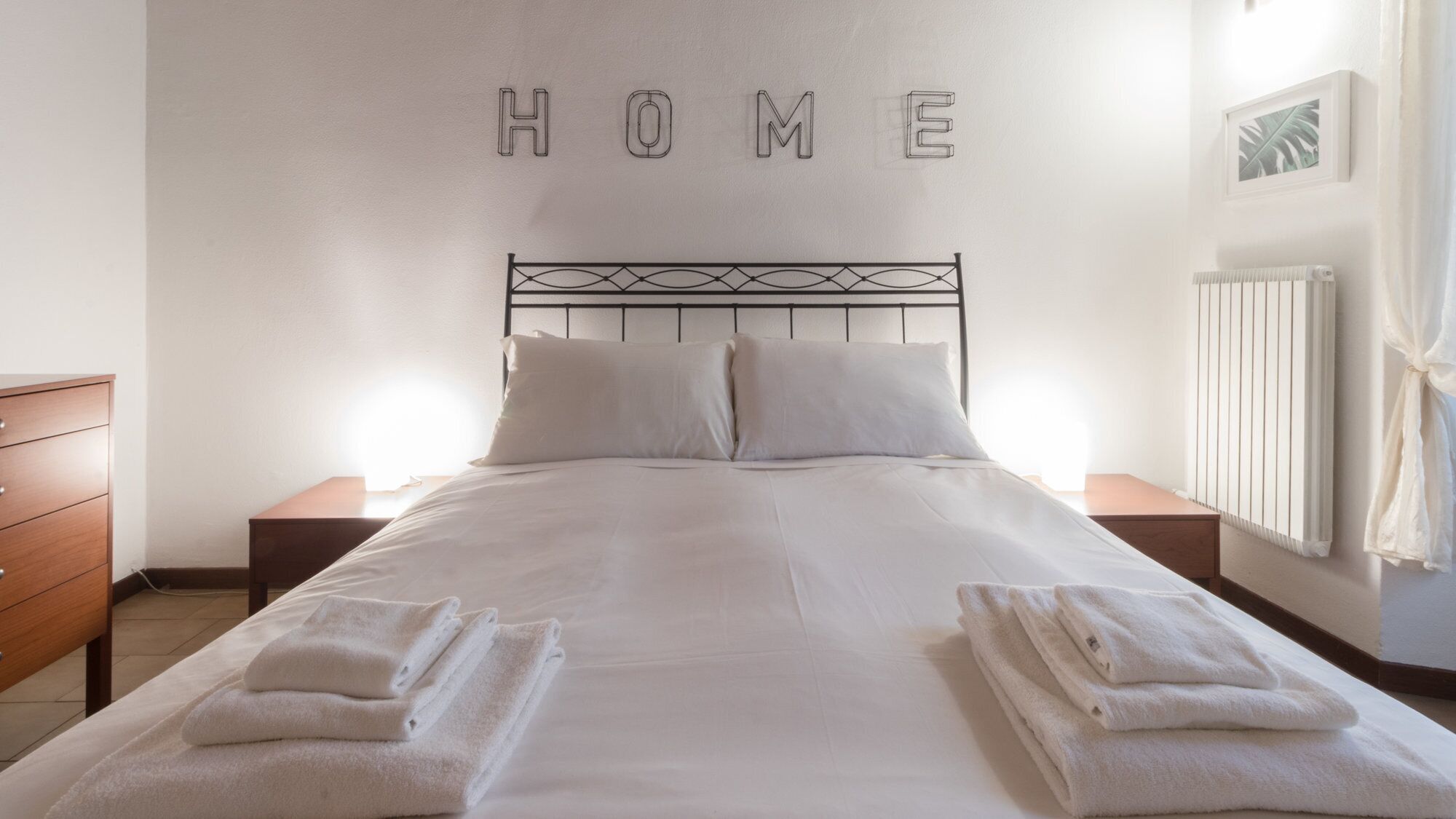 A neatly made bed with folded towels, a headboard, and a wall with the word "HOME" spelled out.