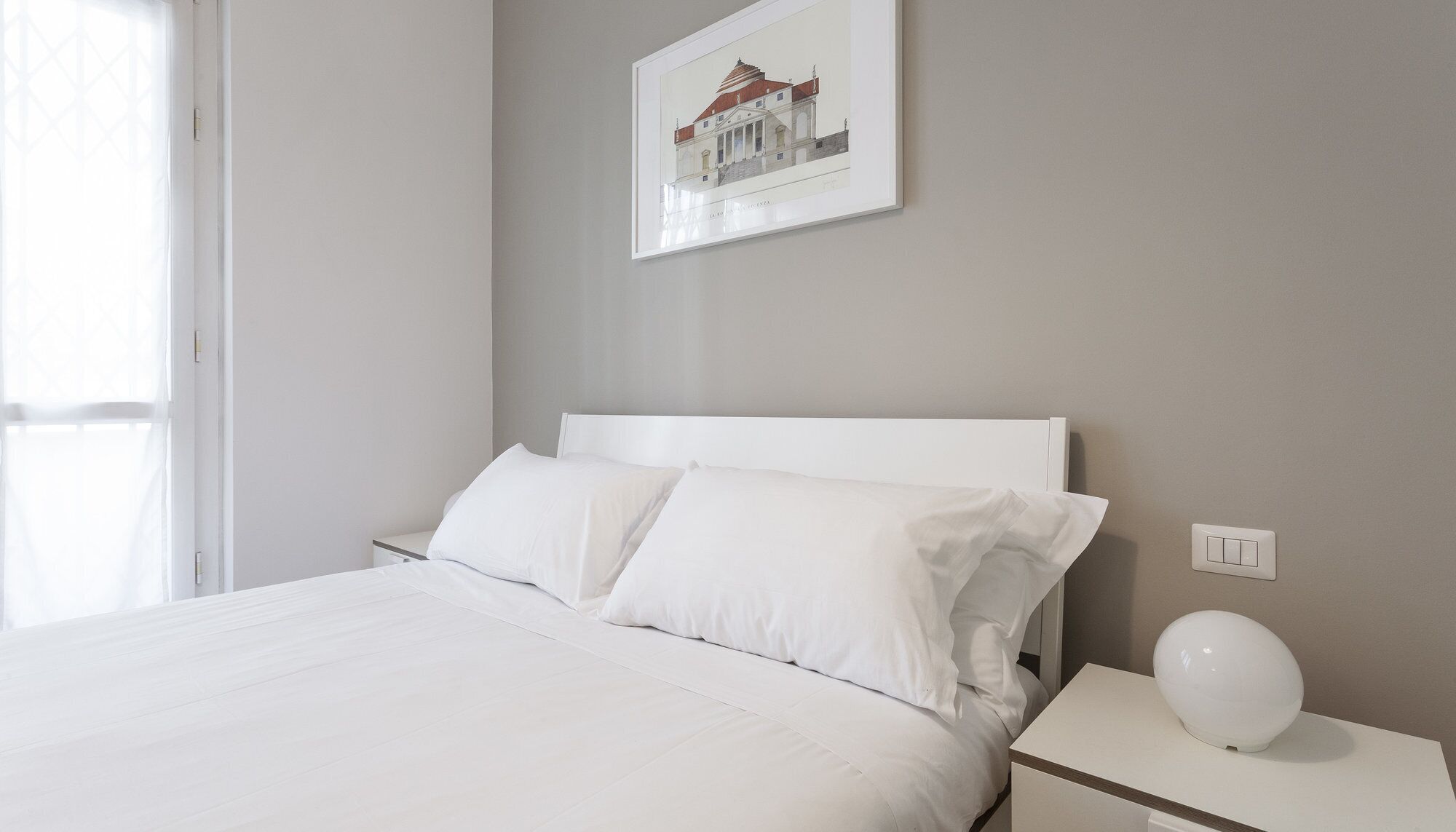Une chambre à coucher au style minimaliste, avec un lit, du linge de lit blanc, une table de chevet avec une lampe et un tableau encadré au mur.
