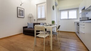 Un espace de vie compact comprenant un coin cuisine, un coin repas avec une table et des chaises, un canapé avec des coussins, un tableau encadré au mur et une fenêtre avec des rideaux.