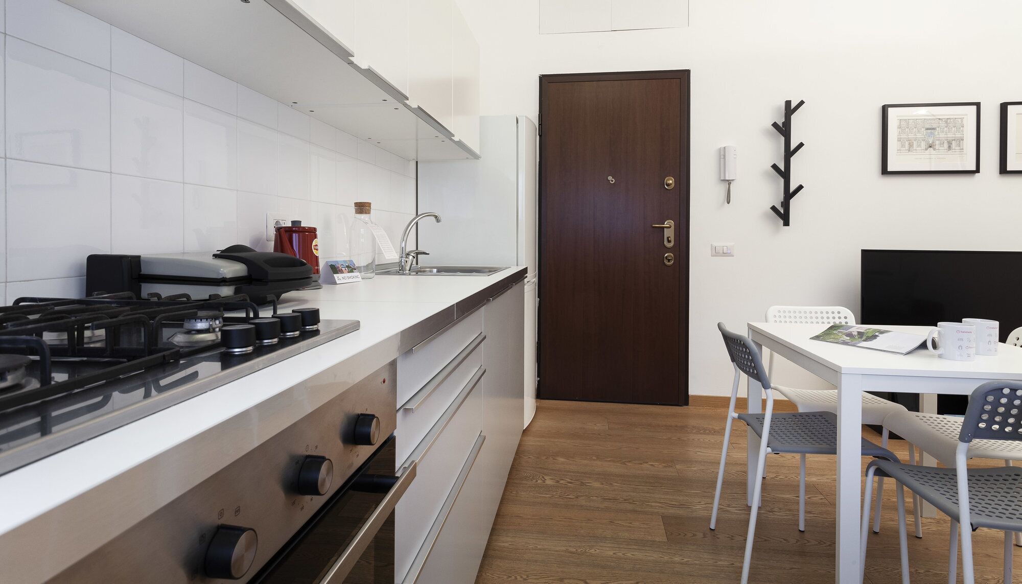A modern kitchen with a gas stove, a sink, and a wooden door. There is a dining table with chairs and a black TV on the wall.