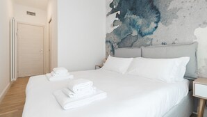 Una cama bien tendida con ropa y toallas blancas, cabecera gris y pared con textura al fondo.