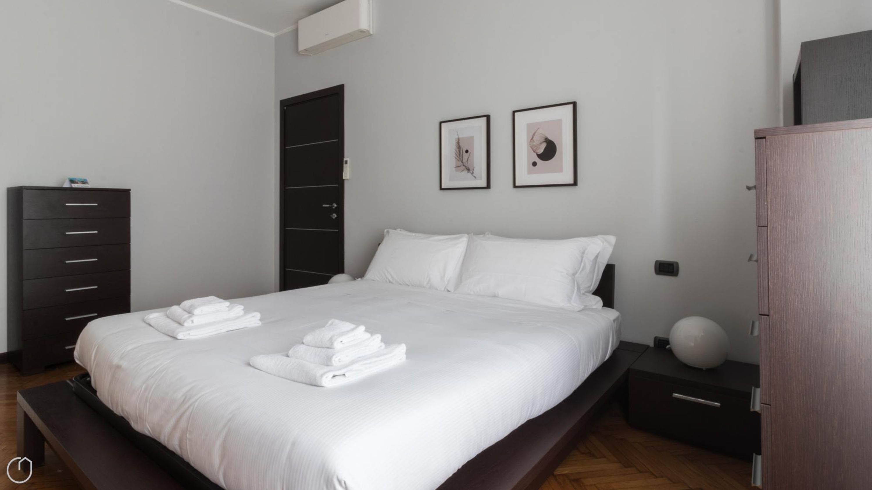 A modern bedroom with a large bed, a dark wardrobe, a bedside table with a lamp, and two framed pictures on the wall.