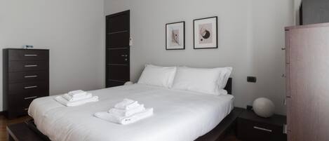 A modern bedroom with a large bed, a dark wardrobe, a bedside table with a lamp, and two framed pictures on the wall.