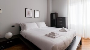 A modern bedroom with a large bed, white linens, a bedside table with a lamp, and a dark dresser. - Italianway Comfort - Antonia Pozzi 5 (Milan)