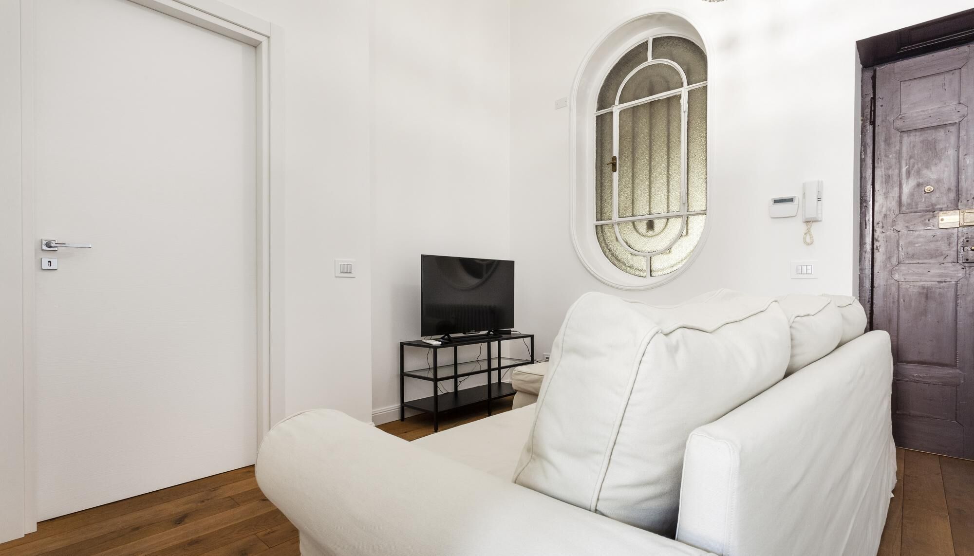 Un salon moderne avec un canapé blanc, un téléviseur posé sur un meuble noir et une porte en bois.