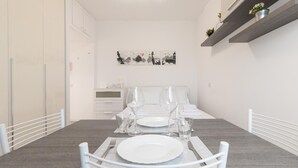 A dining area with a table set for two, white chairs, and a bed with a headboard in the background. - Italianway Easy - Ampere 103 (Milan)