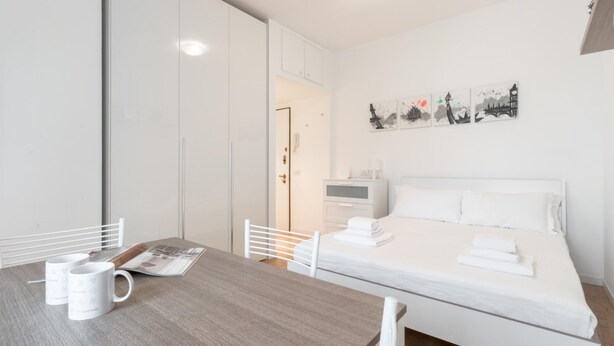 A hotel room with a bed, a desk with a magazine and a mug, a wardrobe, and a wall with artwork. - Italianway Easy - Ampere 103 (Milan)