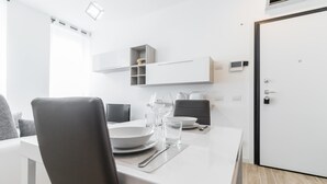 A modern dining area with a white table set for two, a grey chair, and a black chair. There are wine glasses, a bowl, and a plate on the table. A white cabinet with a small shelf is above the table.