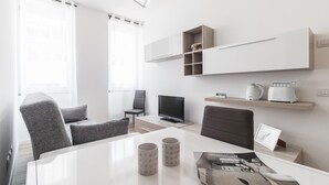 A modern, minimalist living room with a flat-screen TV, a coffee maker, and a toaster on a shelf. There are two grey armchairs and a white dining table with two mugs.