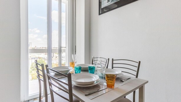 A dining area with a table set for two, chairs, and a view of the city through large windows. - Italianway Comfort - Ripa Ticinese 103 (Milan)